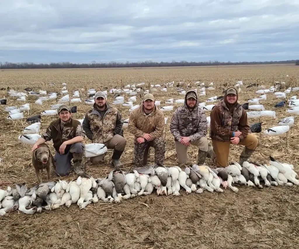 Group of hunters posing with a successful snow goose harvest in a rural field, showcasing a large number of geese after a guided hunt with GETM Waterfowl. The group is surrounded by decoys, capturing the experience of a high-volume conservation hunt.